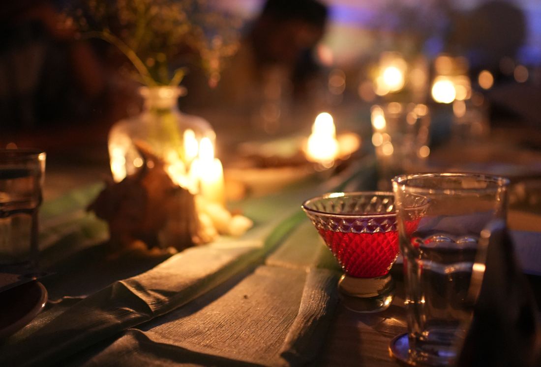 Our Goa, Table No. 02 — the Sol-Kissed Elixir glowing by candlelight against a beach-sunset projection