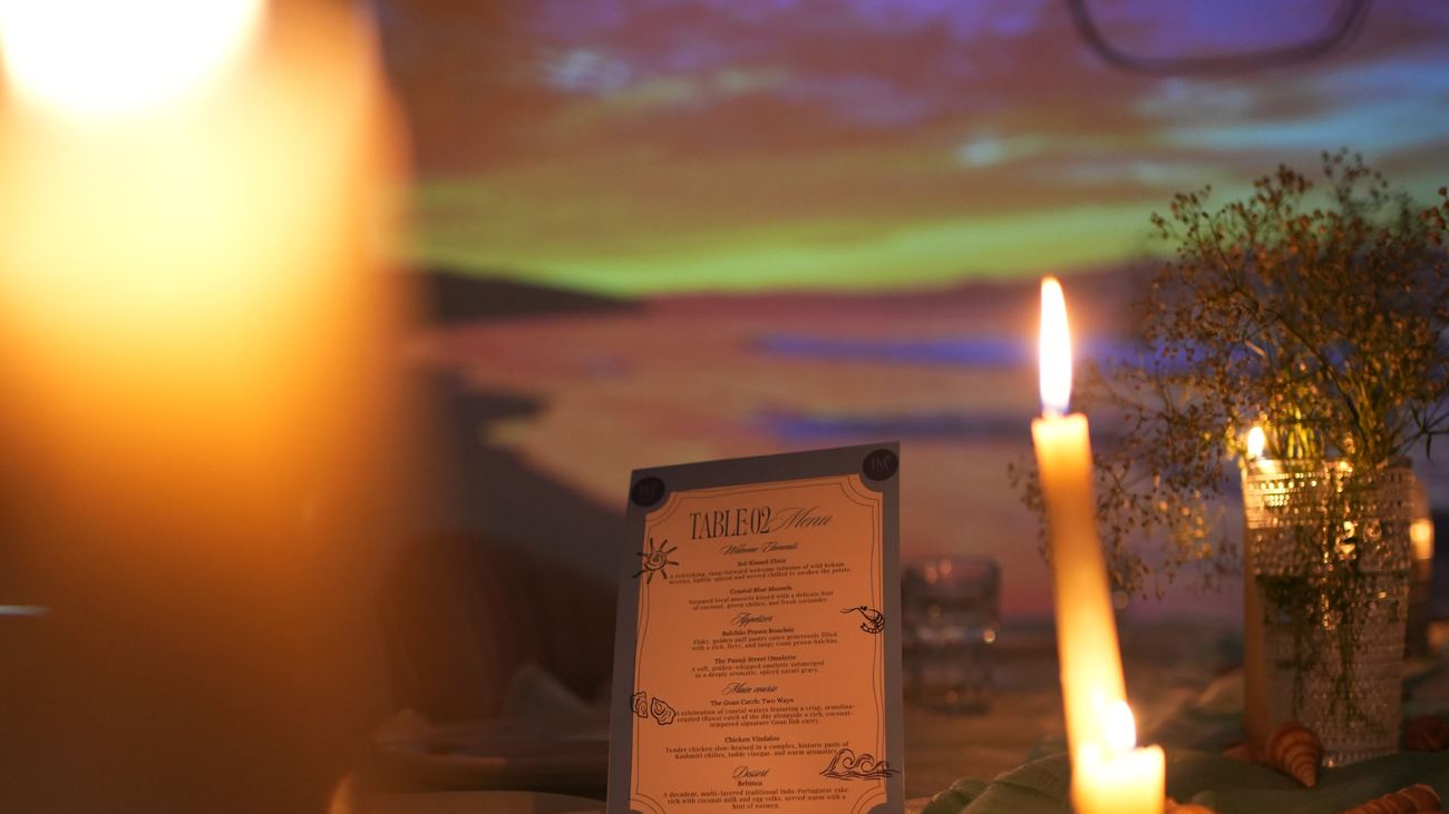 The Table No. 02 menu — Goan courses from sol-kissed elixir to bebinca — lit by candles against a beach-sunset projection